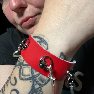 May include: A red leather bracelet with silver metal accents and a small sword charm. The bracelet has a ring on one side and a small ball on the other.