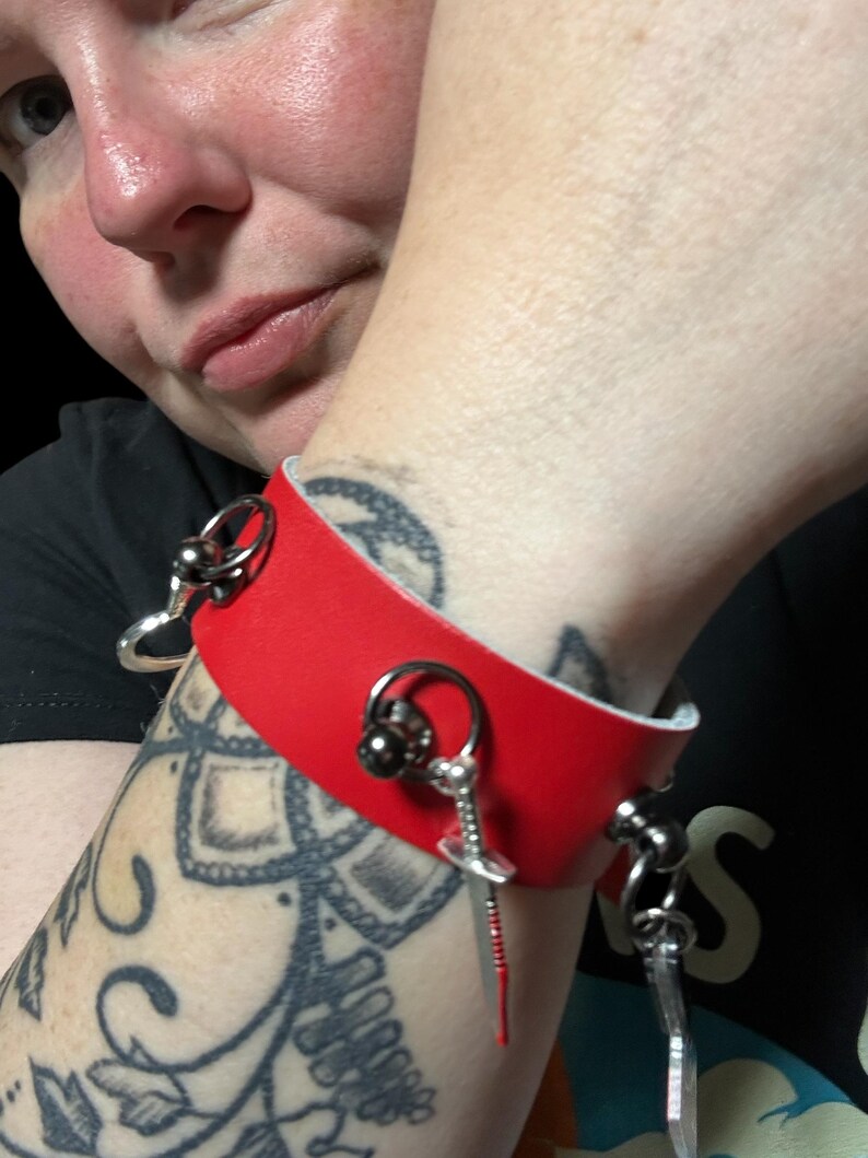 May include: A red leather bracelet with silver metal accents and a small sword charm. The bracelet has a ring on one side and a small ball on the other.