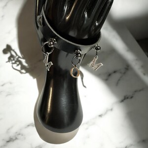May include: Black leather bracelet with silver metal charms. The charms are shaped like the letters 'M' and 'O'.