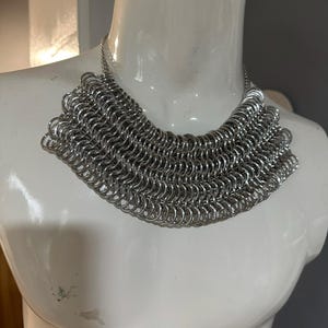 May include: A silver chainmail necklace with a wide, rounded shape. The necklace is made of many small, interconnected metal rings.