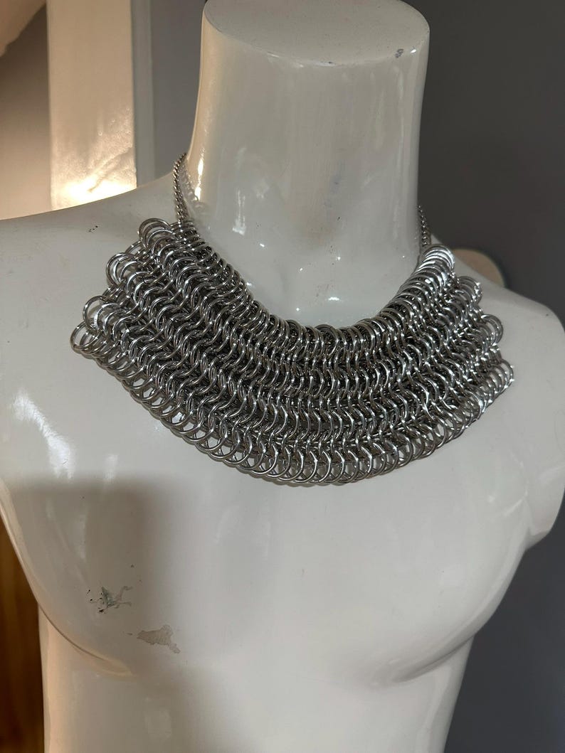 May include: A silver chainmail necklace with a wide, rounded shape. The necklace is made of many small, interconnected metal rings.