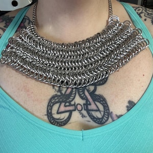 May include: A silver chainmail necklace with a wide, rounded shape. The necklace is made of many small, interconnected metal rings.