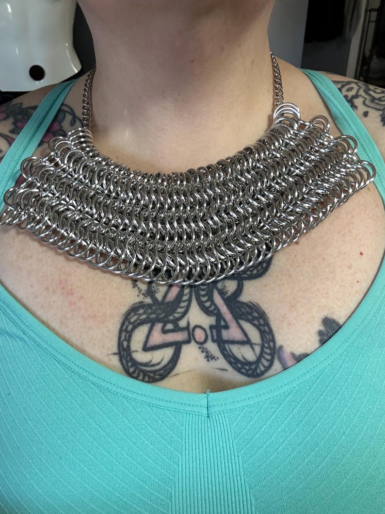 May include: A silver chainmail necklace with a wide, rounded shape. The necklace is made of many small, interconnected metal rings.