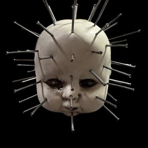 Puede incluir: Una cabeza de muñeca de porcelana con un acabado blanco desgastado, cubierta de numerosos clavos plateados. Los ojos de la muñeca están abiertos y pintados con una expresión realista.