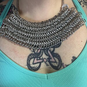 May include: A silver chainmail necklace with a wide, rounded shape. The necklace is made of small, interconnected metal rings.