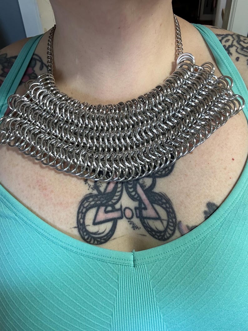 May include: A silver chainmail necklace with a wide, rounded shape. The necklace is made of small, interconnected metal rings.