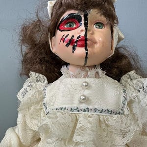 May include: A porcelain doll with brown hair and a white lace dress. The doll's face is painted with a black and red design, creating a spooky and whimsical look.
