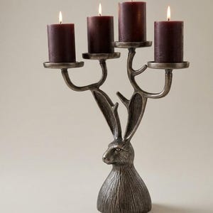 May include: A silver-toned candelabra with a rabbit design, holding four burgundy candles. The candle holders extend from the rabbit's head and body. Matches are placed near the base.