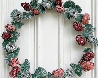 Op de afbeelding: Een decoratieve krans met groene bladeren en bloemknoppen in rood, roze en grijs. De krans hangt aan een witte deur met een messingkleurige haak.