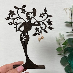 Puede incluir: Un expositor de pendientes de madera con forma de mujer con detalles florales. El expositor es de color marrón oscuro y tiene una base pequeña. Dos pendientes de aro dorados con un diseño floral se muestran en el expositor.