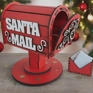 Puede incluir: Un buzón de madera rojo de Santa Mail con las palabras "SANTA MAIL" en blanco. El buzón tiene una puerta abierta y se asienta sobre una base redonda. Un pequeño portae sobres rojo está al lado del buzón. Copos de nieve dorados están esparcidos.