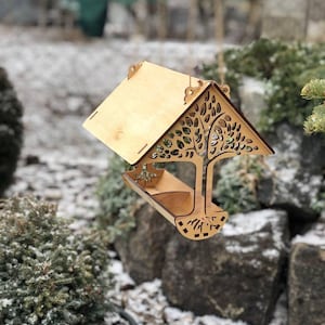 May include: A wooden bird feeder with a tree design cut into the side. The feeder has a pitched roof and a tray for food. The bird feeder is hanging outside in a snowy environment.