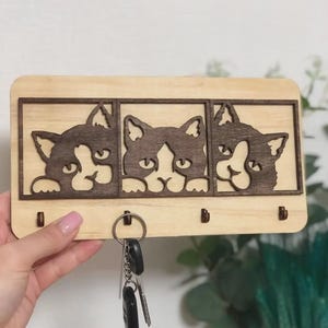 May include: A wooden key holder with three cat silhouettes in individual frames. The cats are dark brown against a light wood. Below the cat faces are three pegs for keys. A set of keys hangs from the key holder, which is made of wood.