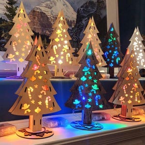 Puede incluir: Decoraciones de árboles de Navidad de madera con formas recortadas de estrellas, bastones de caramelo y hombres de jengibre. Los árboles están iluminados con luces LED de colores, creando una exhibición festiva. Sobre una superficie blanca.
