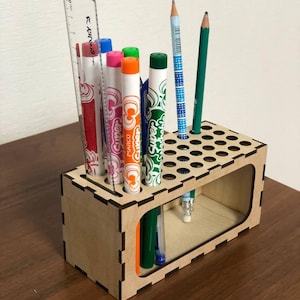 May include: A wooden pencil and pen holder with a clear front panel. The holder has multiple holes for pens and pencils. There are several colored markers, pencils, and a ruler in the holder.