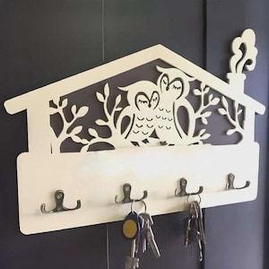May include: A cream-colored wooden key holder shaped like a house, featuring a cut-out design of two owls and foliage. It has five metal hooks for keys, with several keychains hanging from them. The background is a dark gray wall.