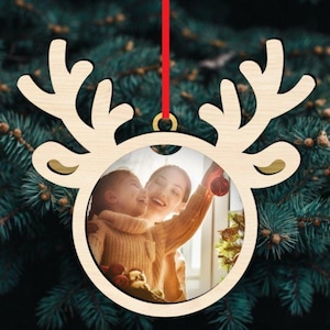 Peut inclure: Ornement de Noël en bois en forme de renne avec un cadre photo circulaire. Le cadre contient une photo d'une femme et d'un enfant. L'ornement est suspendu à un ruban rouge sur fond de sapin de Noël.