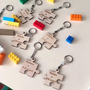 May include: Wooden puzzle piece keychains with silver keyrings. Each keychain has the text "Вітаю 5-Б класі" engraved on it. Colorful toy building blocks in various colors are scattered around the keychains.