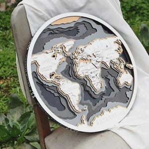 May include: A round, layered wooden world map with a white frame. The map features detailed continents in shades of gray, beige, and white, with country names visible. The map is displayed on a chair with a white cloth.