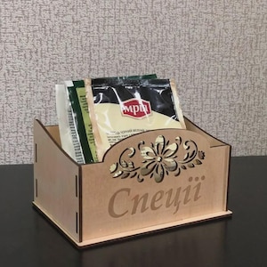 May include: A light brown wooden spice box with a floral design and the word "Спеції" cut out. The box holds several spice packets. The box is on a dark surface.