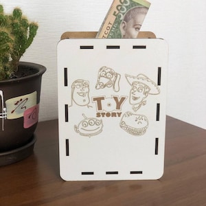 Può includere: Un salvadanaio in legno bianco con immagini incise al laser dei personaggi del film "Toy Story". La scatola ha una fessura nella parte superiore per inserire denaro, con una banconota ucraina da 500 grivnie visibile. La scatola è rettangolare.