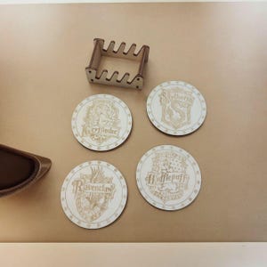 Könnte beinhalten: Set aus vier runden Holz-Untersetzern mit den Wappen der Hogwarts-Häuser: Gryffindor, Slytherin, Hufflepuff und Ravenclaw. Jeder Untersetzer hat ein goldfarbenes Design und ist von einem dekorativen Rand umgeben.