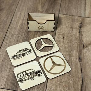 Könnte beinhalten: Vier Untersetzer aus Holz mit einem Mercedes-Benz-Logo und Bildern von Mercedes-Benz-Fahrzeugen. Die Untersetzer befinden sich in einer Holzkiste.