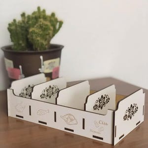 Puede incluir: Un organizador de madera con cuatro compartimentos, con exterior blanco e interior marrón. El panel frontal tiene ilustraciones de alimentos grabadas y las palabras "Sal" y "Pimienta". Los diseños florales adornan los divisores.