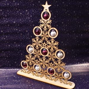 May include: A wooden Christmas tree decoration with snowflake cutouts. The tree holds small, round baubles in red and white. A star tops the tree. The base is light-coloured wood, set against a dark blue background with a star pattern.
