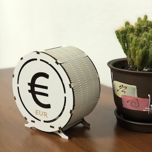 Può includere: Un salvadanaio cilindrico in legno bianco con il simbolo dell'euro e la scritta "EUR". Il design è tagliato al laser con linee parallele. Sullo sfondo è visibile un cactus in un vaso marrone. Il salvadanaio ha una fessura per le monete.