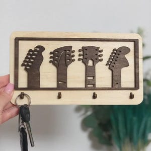 Puede incluir: Llavero de madera con cuatro siluetas de cabezales de guitarra en marrón oscuro sobre un fondo de madera clara. El llavero tiene cuatro ganchos debajo de los cabezales de la guitarra y un juego de llaves colgando.