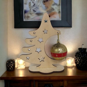Op de afbeelding: Houten kerstboomdecoratie met stervormige uitsparingen en een goud-rode kerstbal eraan. De bal is rond en heeft gouden details. Het geheel staat op een houten voet.