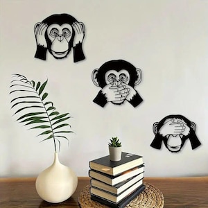 Può includere: Arte da parete in metallo nero con tre facce di scimmia nelle pose 'non sentire il male, non vedere il male, non dire il male'. Le scimmie sono montate su una parete bianca. Un vaso bianco con una pianta verde e una pila di libri sono in primo piano.