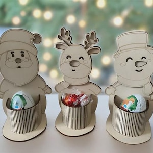 Puede incluir: Decoraciones navideñas de madera con un Papá Noel, un reno y un muñeco de nieve. Cada figura tiene una cesta para guardar golosinas. La figura de Papá Noel sostiene un huevo, el reno caramelos envueltos y el muñeco de nieve un huevo.