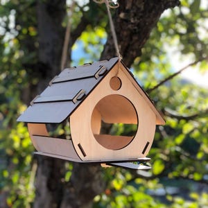 Pode incluir: Um comedouro para pássaros de madeira em forma de casa com uma abertura arredondada. O comedouro está pendurado em uma corda e é feito de madeira marrom clara.