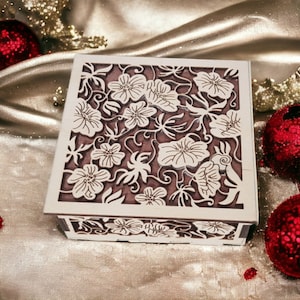 May include: A square wooden box with a floral design carved into the lid. The box is painted white with a brown interior. The floral pattern is intricate and detailed. The box is sitting on a gold satin surface with red Christmas ornaments.