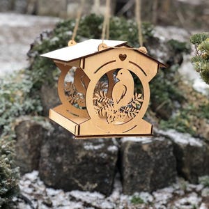 May include: A hanging wooden bird feeder with a roof and a heart-shaped cutout. The feeder features a bird silhouette and leaf design within a circular frame. The natural wood colour contrasts with the snowy background.