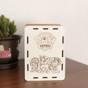 Può includere: Scatola di legno bianca con un bordo marrone e un logo di Paw Patrol. Il logo è un'impronta di zampa bianca all'interno di un cerchio con le parole "PAW PATROL" sotto. Ci sono tre cani dei cartoni animati dello show Paw Patrol sulla scatola.