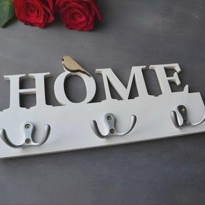 以下が含まれることがあります： 「HOME」の文字が大きく書かれた白い木製の壁掛けフック。3つのシルバーのフックと、文字「O」の上に小さな木製の鳥がいます。背景には赤いバラがあります。