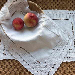 May include: Two fresh peaches resting on a white linen napkin with intricate cutwork embroidery and a scalloped edge. The napkin is draped over a woven basket, and a matching rectangular placemat with similar lace detailing lies underneath.