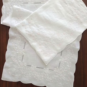 May include: Two white rectangular linens featuring intricate white floral embroidery and delicate cutwork details along the borders. The edges are finished with a scalloped design, and the fabric has a semi-sheer, lightweight appearance.