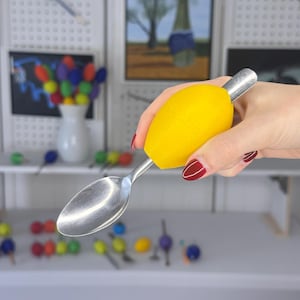 May include: A stainless steel spoon with a bright yellow, ergonomic handle. The handle is designed for easy gripping. The spoon is held in a hand with red nail polish. The background includes artwork and decorative items.