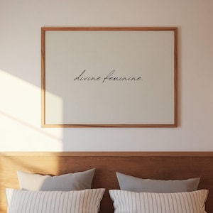 May include: A framed print with the words "divine feminine" in elegant script. The artwork is set against a neutral background and framed in a light brown wooden frame. The print hangs above a bed with gray pillows.
