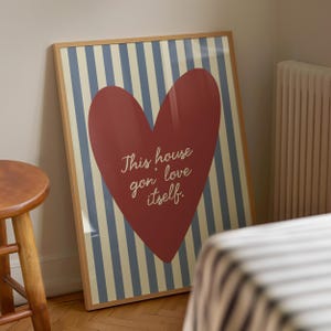 Könnte beinhalten: Gerahmter Kunstdruck mit einem großen roten Herz auf einem Hintergrund aus blauen und cremefarbenen Streifen. Das Herz trägt den Text "This house gon love itself". Der Rahmen ist hellbraun.
