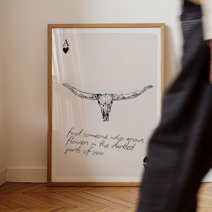 May include: A framed art print with a black and white bull skull illustration and the text "find someone who grows flowers in the darkest parts of you". The Ace of Hearts symbol is also visible.