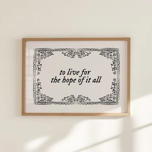 May include: A framed print with the words "to live for the hope of it all" in black script. The text is centered on a white background, framed by a decorative black floral border. The frame is a light brown color.