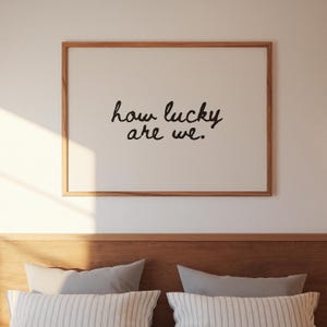 May include: A framed print with the words "how lucky are we" in black script. The print has a light background and a natural wood frame. The image is above a bed with pillows.