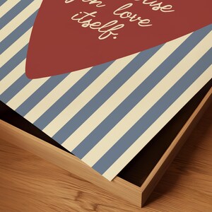 May include: A framed print featuring a red heart shape and blue and cream striped background. The print includes the white text "because love itself." The frame is made of light-colored wood.