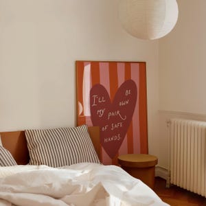Pu&ograve; includere: Una scena di camera da letto con un letto con lenzuola bianche, cuscini a righe e una grande opera d'arte incorniciata. L'opera d'arte ha uno sfondo a righe rosse e rosa con una forma di cuore e il testo "I'll be my own pair of safe hands."