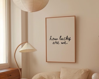 May include: A framed print with the text "how lucky are we." hangs on a beige wall above a bed with striped bedding. A paper lantern and a curved wooden lamp with a pleated lampshade are also visible.